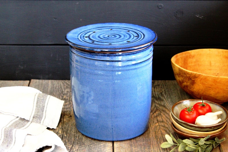 Puede incluir: Un contenedor de compost de cer&aacute;mica azul con una tapa que presenta un dise&ntilde;o en espiral y agujeros de drenaje. El contenedor est&aacute; sobre una superficie de madera.