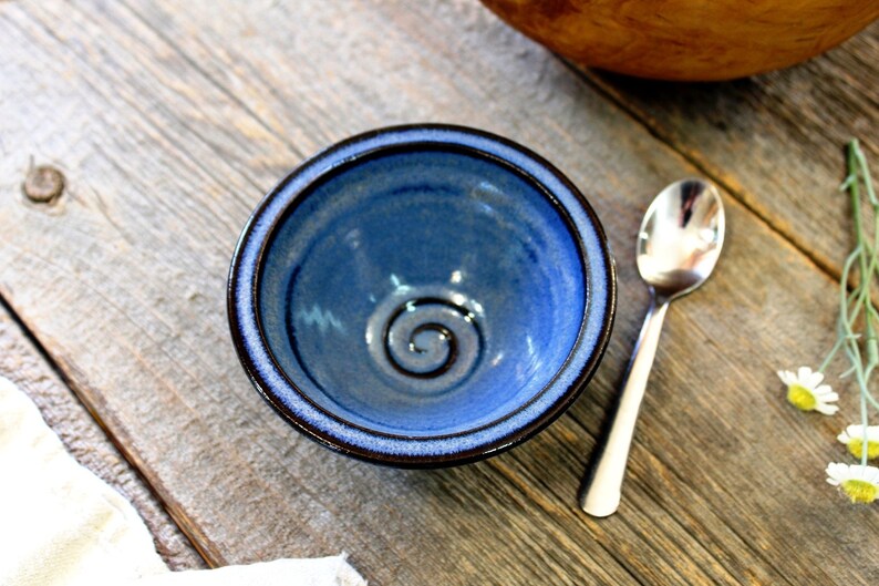 May include: A handcrafted blue ceramic bowl with a spiral pattern in the centre. The bowl has a dark blue rim and a lighter blue interior. A silver spoon sits beside the bowl on a wooden surface.