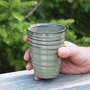 May include: A green ceramic mug with brown stripes, held in a hand against a blurred green background.