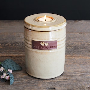 May include: A beige ceramic candle holder with a lid, featuring a burning tea light. The holder has horizontal ridges and a brown rectangular label with two hearts and the words "I love, my love."