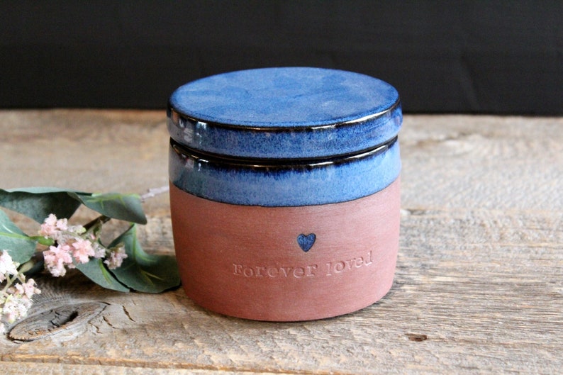 May include: A blue and brown ceramic jar with a lid. The jar has the words "Forever loved" and a small heart engraved on the side.