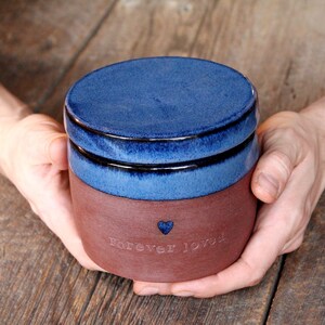 May include: A blue and brown ceramic jar with a lid. The jar has the words "Forever Loved" and a small heart etched into the side.