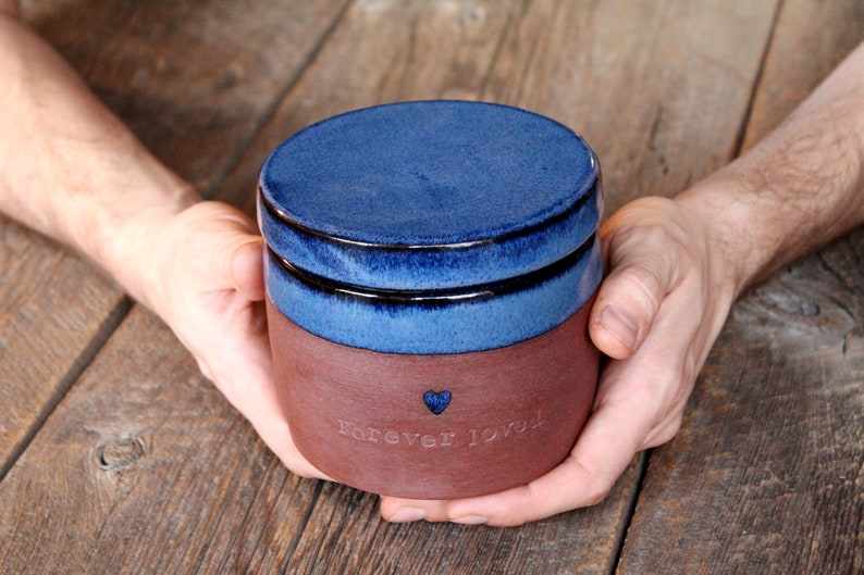 May include: A blue and brown ceramic jar with a lid. The jar has the words "Forever Loved" and a small heart etched into the side.