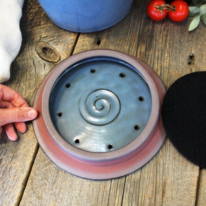 Puede incluir: Una tapa de compostador de cer&aacute;mica con un esmalte azul y un dise&ntilde;o en espiral. La tapa tiene una serie de agujeros para el drenaje y la ventilaci&oacute;n. Un filtro de carb&oacute;n vegetal negro se muestra junto a la tapa.