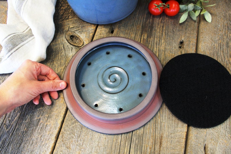 Puede incluir: Una tapa de compostador de cer&aacute;mica con un esmalte azul y un dise&ntilde;o en espiral. La tapa tiene una serie de agujeros para el drenaje y la ventilaci&oacute;n. Un filtro de carb&oacute;n vegetal negro se muestra junto a la tapa.