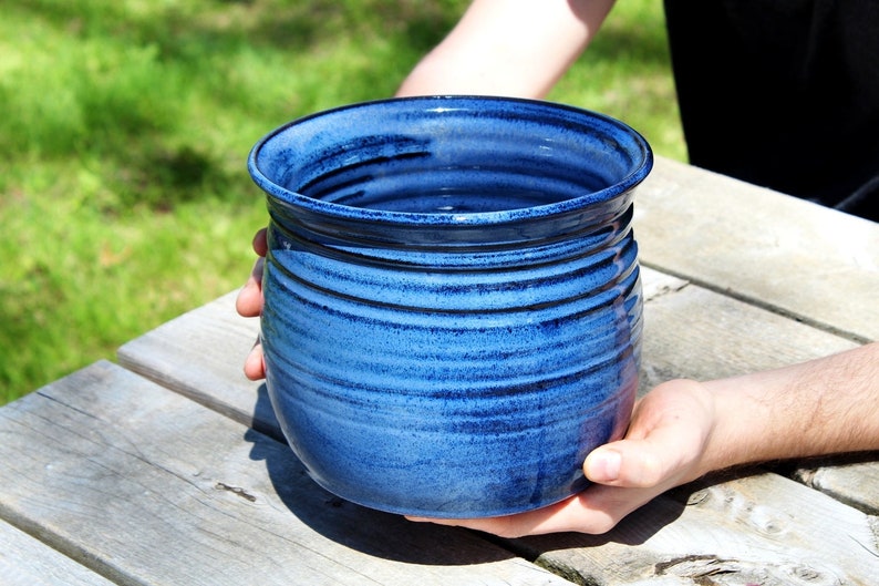 Może przedstawiać: Duży, ciemnoniebieski ceramiczny garnek o zaokrąglonym kształcie i lekko rozszerzającym się brzegu. Garnek ma fakturowaną, lekko nakrapianą glazurę i jest wykonany ręcznie.