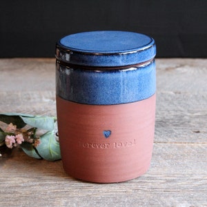 May include: A blue and terracotta ceramic jar with a lid. The jar has a small heart and the words "forever loved" inscribed on the side.
