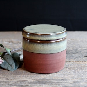 Peut inclure: Un petit pot rond en céramique avec un couvercle. Le pot est émaillé d'une couleur vert clair et marron, avec une base en argile rouge. Le couvercle est également émaillé d'une couleur vert clair et marron.