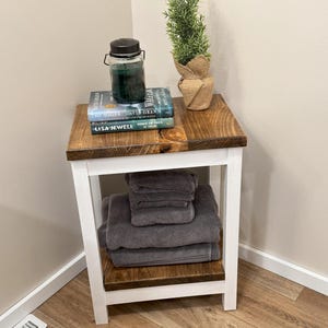 May include: White wooden side table with two shelves. The top shelf has a green candle in a glass jar, a potted green plant, and three books stacked on top of each other. The bottom shelf has a stack of gray towels. The table is in a room with light brown wood floors.