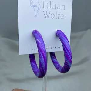Peut inclure: Boucles d'oreilles créoles en forme de spirale violette et blanche sur une carte blanche avec le texte "Lillian Wolfe handmade jewelry".