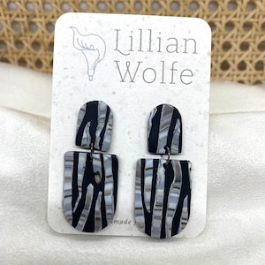 Puede incluir: Un par de pendientes colgantes de rayas blancas y negras con una pequeña pieza superior negra. Los pendientes están en una tarjeta blanca con el texto "Lillian Wolfe" impreso en ella.
