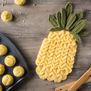 May include: A yellow crocheted pineapple pot holder with green crocheted leaves. The pot holder is on a wooden surface with a baking sheet of muffins.