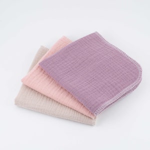 May include: Three cotton muslin cloths in shades of beige, pink, and purple. The cloths are folded and stacked on top of each other.