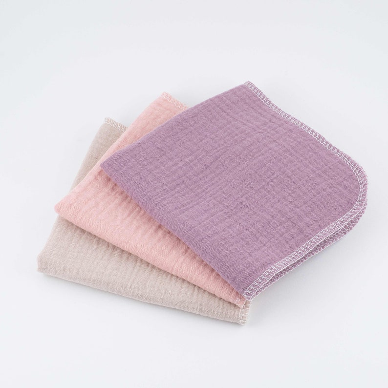 May include: Three cotton muslin cloths in shades of beige, pink, and purple. The cloths are folded and stacked on top of each other.