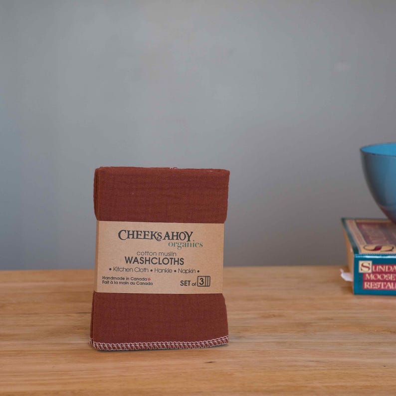 May include: A set of three brown cotton muslin washcloths, folded and stacked. The label reads "Cheeks Ahoy Organics Cotton Muslin Washcloths Kitchen Cloth Hankie Napkin Set of 3 Handmade in Canada Fait &agrave; la main au Canada".