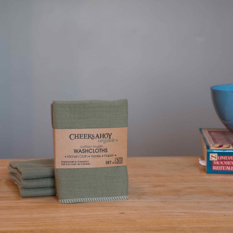 May include: A set of three green cotton muslin washcloths, folded and stacked on a wooden surface. The washcloths are labeled "CHEEKS AHOY. organics cotton muslin WASHCLOTHS Kitchen Cloth Handker Napkin SET of 3 Handmade in Canada Fait &agrave; la main au Canada."