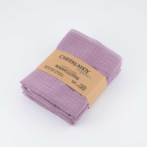 May include: A set of three purple cotton muslin washcloths, handmade in Canada. The washcloths are folded and stacked, with a brown label that reads "CHEEKS AHOY organics cotton muslin WASHCLOTHS SET of 3".