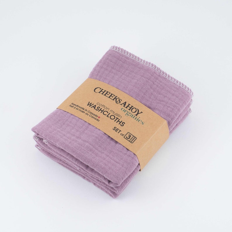 May include: A set of three purple cotton muslin washcloths, handmade in Canada. The washcloths are folded and stacked, with a brown label that reads "CHEEKS AHOY organics cotton muslin WASHCLOTHS SET of 3".