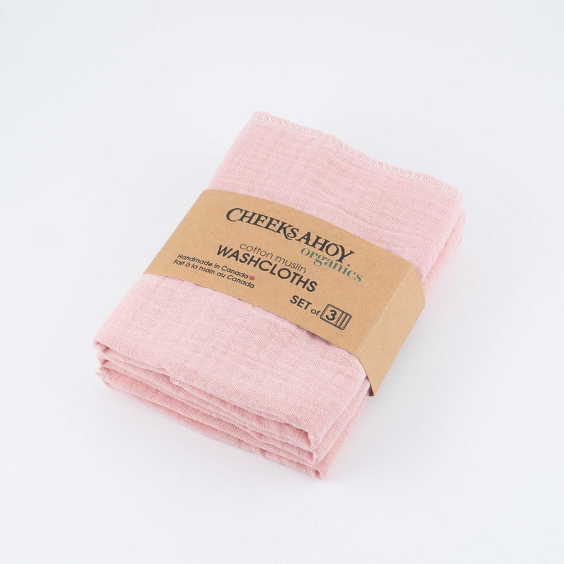 May include: A set of three pink cotton muslin washcloths, folded and packaged with a brown label that reads "CHEEKS AHOY organics cotton muslin WASHCLOTHS SET of 3".