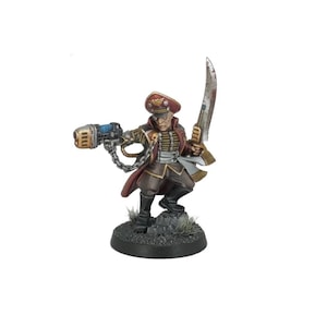 May include: Miniature figurine of a military officer in a red and brown uniform, holding a large energy weapon and a sword. The figure is on a circular base with grass and rocks. The officer wears a peaked cap and has a stern expression.