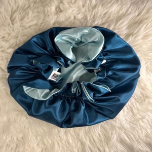 Puede incluir: Un gorro reversible de satén en color verde azulado y azul claro. El gorro tiene un borde fruncido con un cordón. El interior es de satén azul claro y el exterior es de satén verde azulado. Una pequeña etiqueta con la palabra "MARIE" es visible.