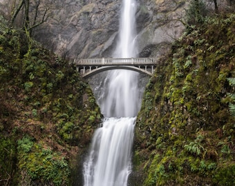 May include: A scenic waterfall cascades down a rocky cliff, flowing under a stone bridge. Lush green vegetation covers the cliff sides, creating a natural frame around the waterfall. The bridge has arched supports.