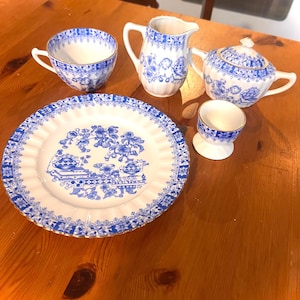 May include: A white porcelain tea set featuring a blue floral and architectural pattern with gold trim. The set includes a scalloped plate, a teacup, a creamer pitcher, a lidded sugar bowl with two handles, and a small egg cup, all matching.