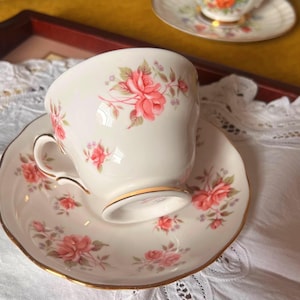 Peut inclure: Un ensemble tasse et soucoupe en porcelaine blanche orné d'un motif floral de roses roses délicates avec des feuilles vertes et un liseré doré sur les bords. Un deuxième ensemble assorti est visible en arrière-plan, légèrement flou.