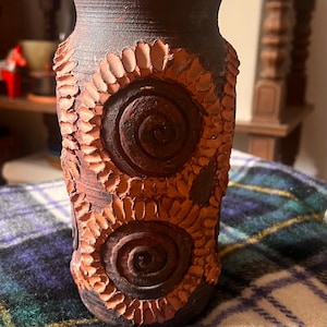 May include: A cylindrical dark brown clay vase featuring two prominent circular spiral motifs on the front. The spirals are surrounded by textured, petal-like indentations in a lighter terracotta colour, creating a tactile, relief-style surface.