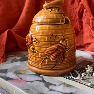 May include: A ceramic honey pot shaped like a beehive, with a lid and a small handle. The pot is tan with embossed bees and a textured surface. A decorative kitchen item.