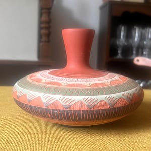 May include: A low-profile terracotta vase with a narrow neck and a wide, rounded body. The surface features intricate, carved geometric patterns in shades of muted green, white, and dark brown, contrasting against the natural reddish-orange clay.