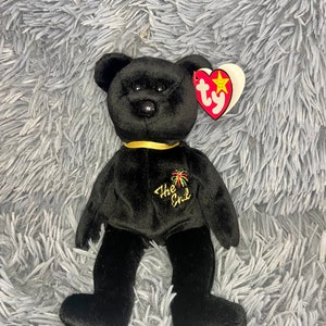 Puede incluir: Oso de peluche negro Beanie Baby con una cinta dorada alrededor del cuello. El oso tiene una etiqueta bordada negra que dice "The End" con un diseño de fuegos artificiales. El oso está sentado sobre una superficie gris y peluda.