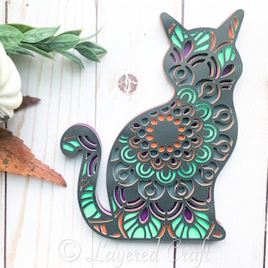 Puede incluir: Una silueta de gato negro con intrincados diseños de mandala en tonos de verde, morado y naranja. El gato está sentado con la cola enroscada alrededor de su cuerpo.
