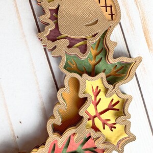 May include: Layered paper craft featuring an acorn, leaves, and branches in shades of brown, green, red, and yellow. The craft is made with textured paper and has a layered effect.