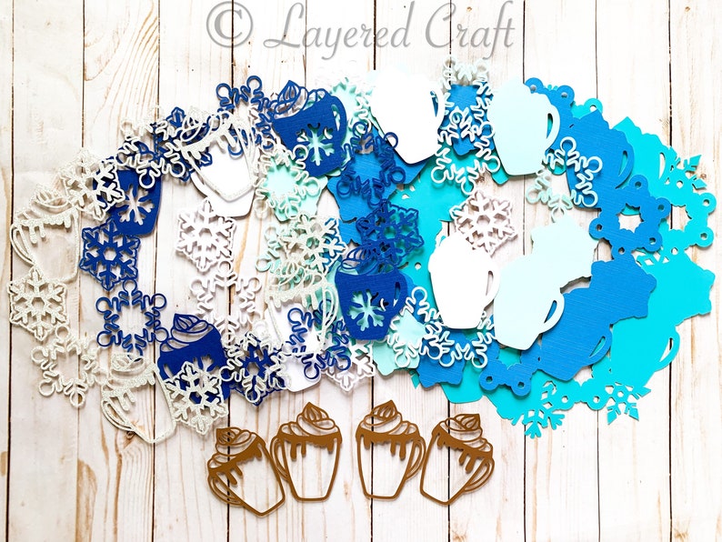 May include: A collection of die-cut paper shapes in winter and holiday themes. Includes snowflakes, mugs, and hot chocolate cups in shades of blue, white, and brown. The Layered Craft logo is visible in the upper right corner.