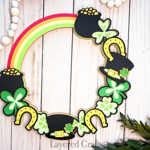 May include: A St. Patrick's Day wreath with a rainbow, pots of gold, shamrocks, and horseshoes. The wreath is made of paper and is decorated with green, gold, and black colors.