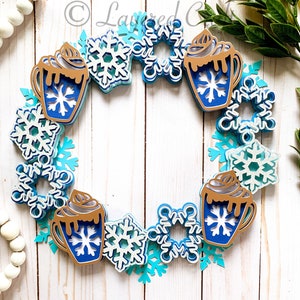 May include: A winter-themed wreath featuring layered paper cutouts. The wreath is adorned with blue and white snowflakes and hot cocoa mugs with whipped cream. The color palette includes shades of blue, white, and brown.