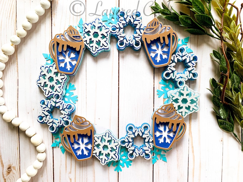 May include: A winter-themed wreath featuring layered paper cutouts. The wreath is adorned with blue and white snowflakes and hot cocoa mugs with whipped cream. The color palette includes shades of blue, white, and brown.