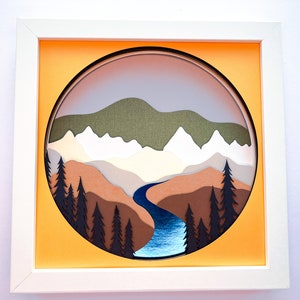 May include: A framed paper art piece featuring a layered landscape design. The artwork showcases a mountain range, a river, and trees in shades of green, brown, and blue, set against an orange background within a white frame.