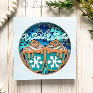 Puede incluir: Una tarjeta de papel azul y marrón con un círculo recortado en el centro. El círculo presenta dos tazas de chocolate caliente con crema batida y copos de nieve. El texto "Warm Wishes" está escrito en una fuente cursiva sobre las tazas.