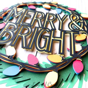 Pu&ograve; includere: Una ghirlanda natalizia di carta a strati con le parole "MERRY & BRIGHT" in nero e oro. La ghirlanda &egrave; decorata con luci colorate rosse, gialle, blu e rosa e foglie di carta verdi.