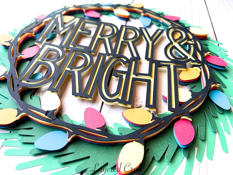 Pu&ograve; includere: Una ghirlanda natalizia di carta a strati con le parole "MERRY & BRIGHT" in nero e oro. La ghirlanda &egrave; decorata con luci colorate rosse, gialle, blu e rosa e foglie di carta verdi.