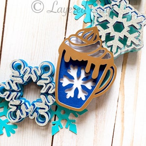 May include: A layered paper art piece featuring a hot chocolate mug and snowflakes in shades of blue, silver, and gold. The mug has a snowflake cutout and whipped cream. The snowflakes are in various sizes and designs, creating a winter theme.