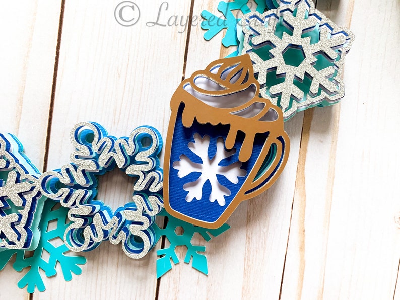 May include: A layered paper art piece featuring a hot chocolate mug and snowflakes in shades of blue, silver, and gold. The mug has a snowflake cutout and whipped cream. The snowflakes are in various sizes and designs, creating a winter theme.