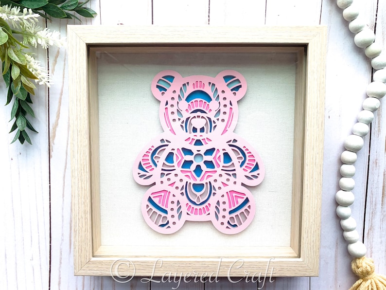 May include: A pink and blue layered paper bear cutout in a wooden frame. The bear is decorated with intricate mandala patterns. The frame has a white background and the text "Layered Craft" is visible in the bottom right corner.