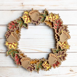 May include: A fall-themed wreath made of layered paper leaves in shades of red, orange, yellow, and green. The wreath is decorated with brown acorns with a cross-stitch pattern.