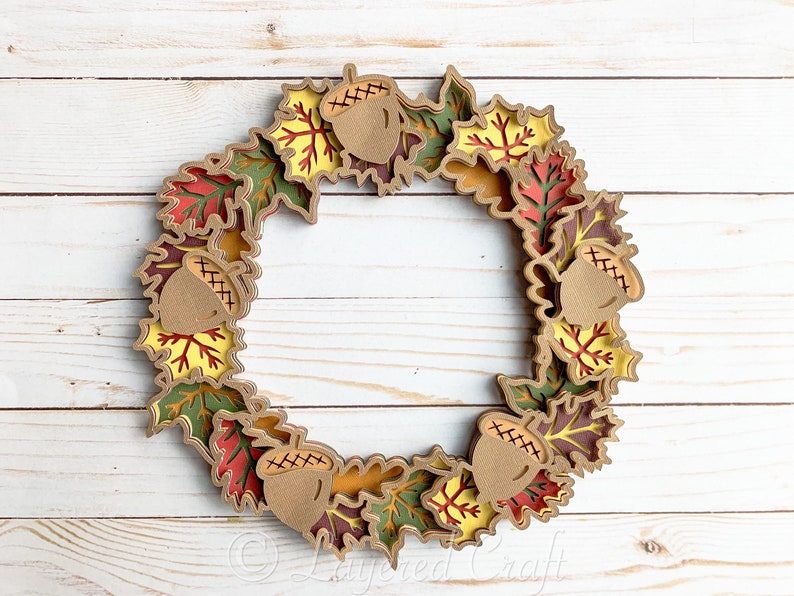 May include: A fall-themed wreath made of layered paper leaves in shades of red, orange, yellow, and green. The wreath is decorated with brown acorns with a cross-stitch pattern.