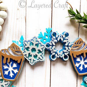 May include: A layered paper craft winter-themed garland featuring blue and silver snowflakes and hot cocoa mugs. The mugs have brown whipped cream and blue snowflake designs. The garland is set against a white wooden background.