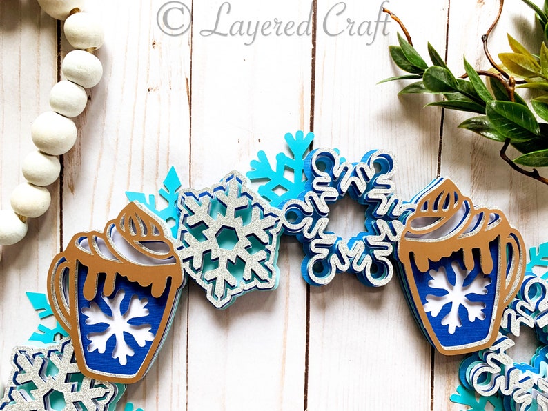 May include: A layered paper craft winter-themed garland featuring blue and silver snowflakes and hot cocoa mugs. The mugs have brown whipped cream and blue snowflake designs. The garland is set against a white wooden background.