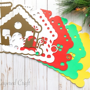 Op de afbeelding: Vijf gelaagde papieren gingerbread house uitsnijdingen in verschillende kleuren, waaronder bruin, wit, rood, groen en geel. De uitsnijdingen zijn in een waaier op een wit houten oppervlak gerangschikt. De uitsnijdingen hebben ingewikkelde details, waaronder snoepstokken, ramen en een schoorsteen.